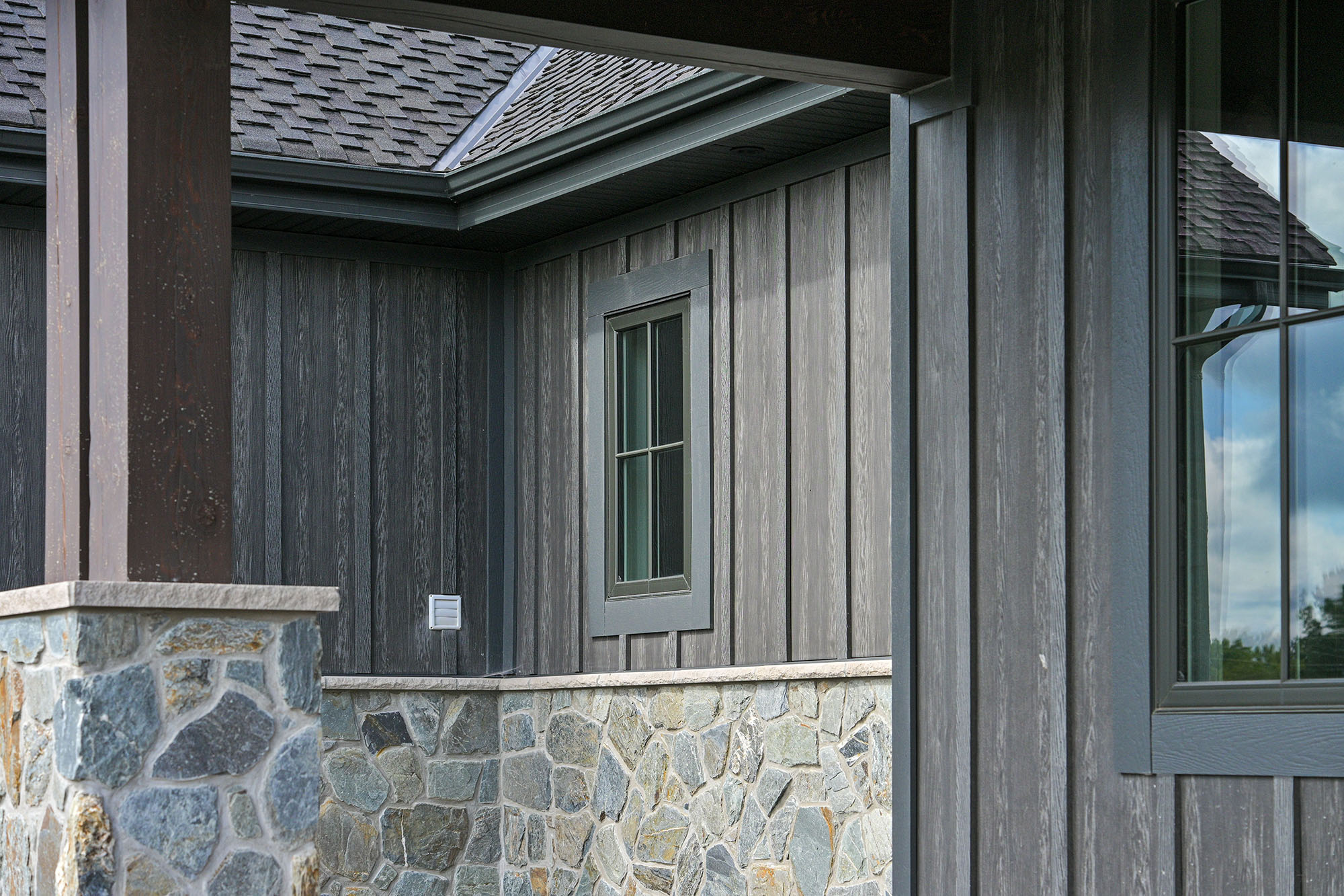 Close-up of grey board-and-batten siding, stone veneer, asphalt shingles, soffit and fascia on a home exterior.