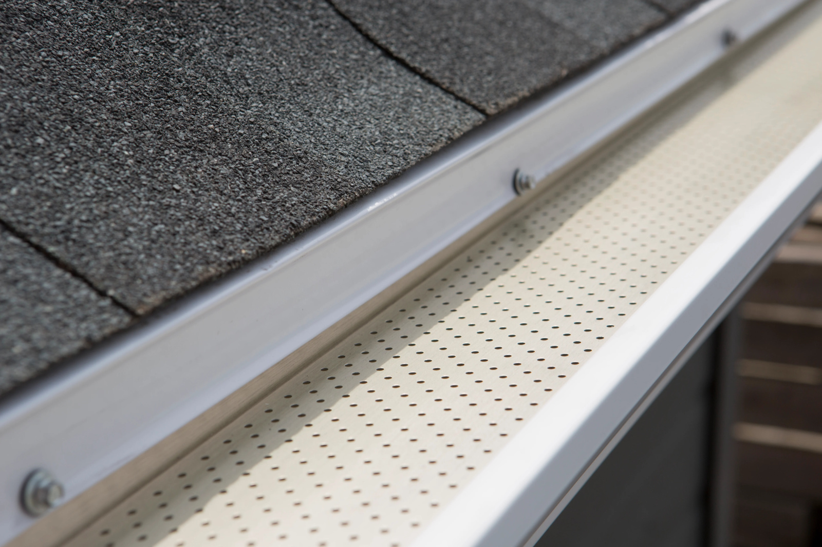 Close-up of perforated aluminum gutter guard installed along an eavestrough beside asphalt shingles.