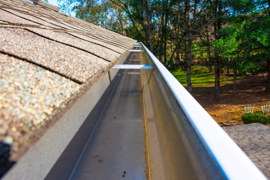 Clean aluminum eavestrough beside weathered asphalt shingles, backyard trees in the background.