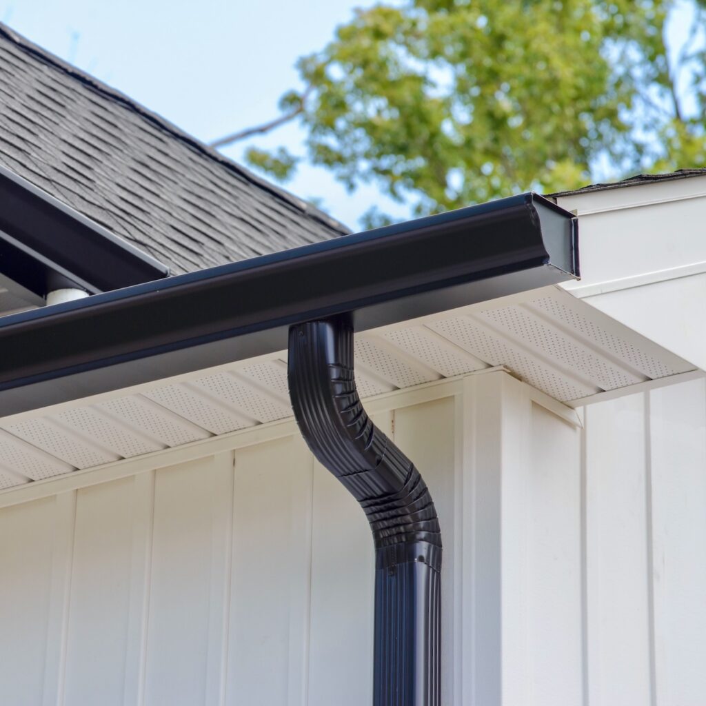 Black eavestrough and downspout on white board-and-batten siding with vented soffit.