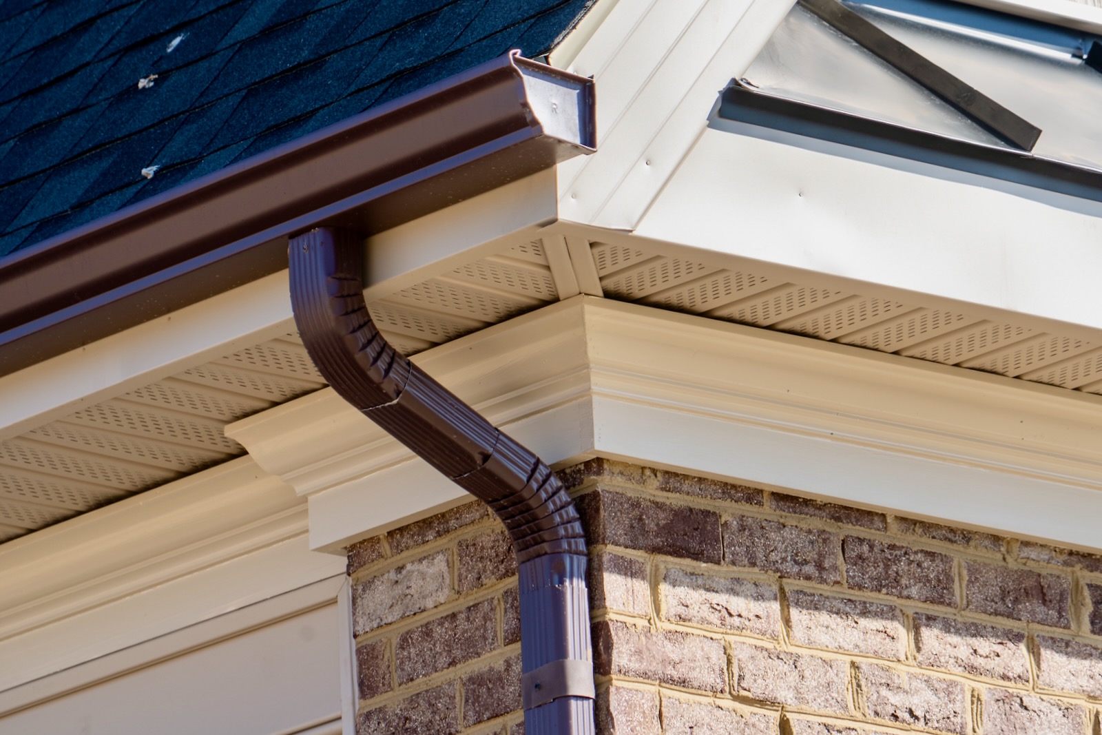 Brown metal downspout elbow against beige soffit, fascia and brick wall under dark shingles.