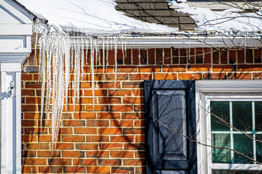 Long icicles hanging from an eavestrough and ice dam along a snowy roof edge on a brick house.