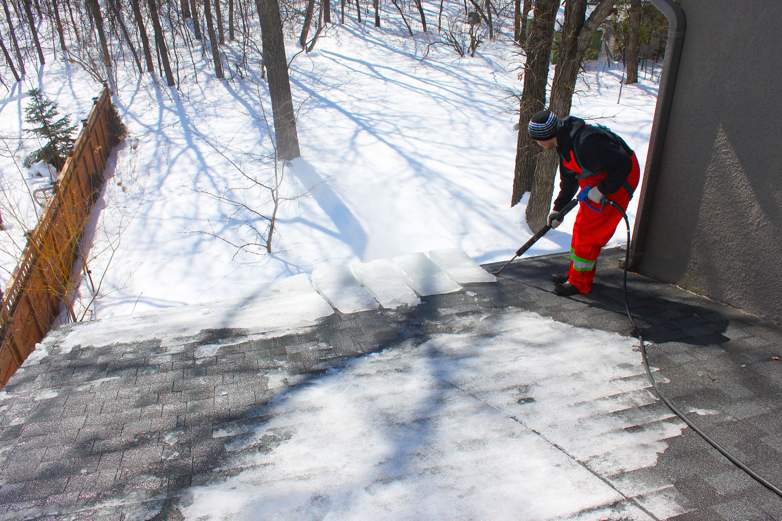 Worker in red coveralls steaming ice and snow off a residential asphalt shingle roof in winter.