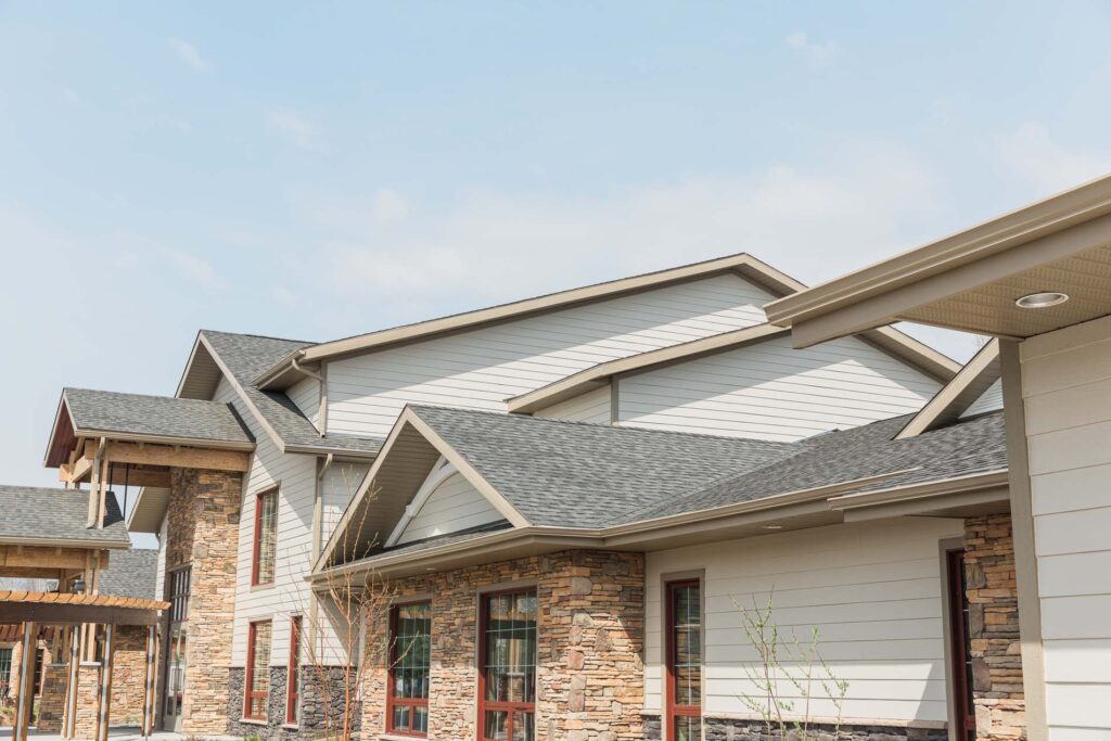 Residential building exterior with light siding, stone accents, grey shingle roof, and covered entryway.