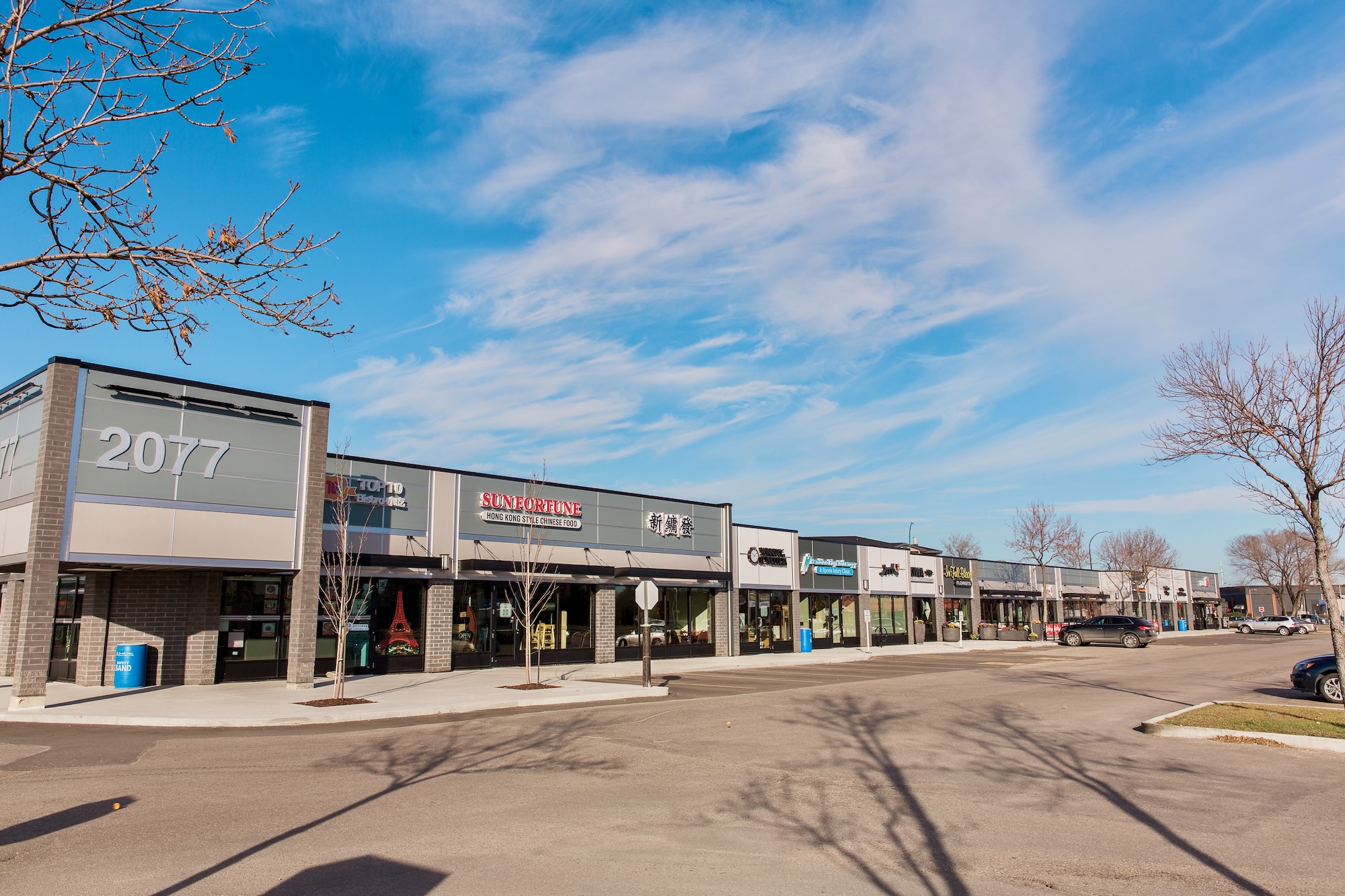 2077Pembina Strip Mall showing half a dozen store fronts, with a parking lot and some shadows from trees.