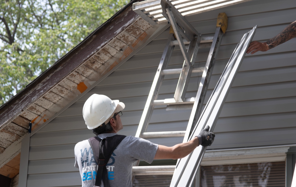 Roofer passes a metal siding panel to a coworker beside a ladder